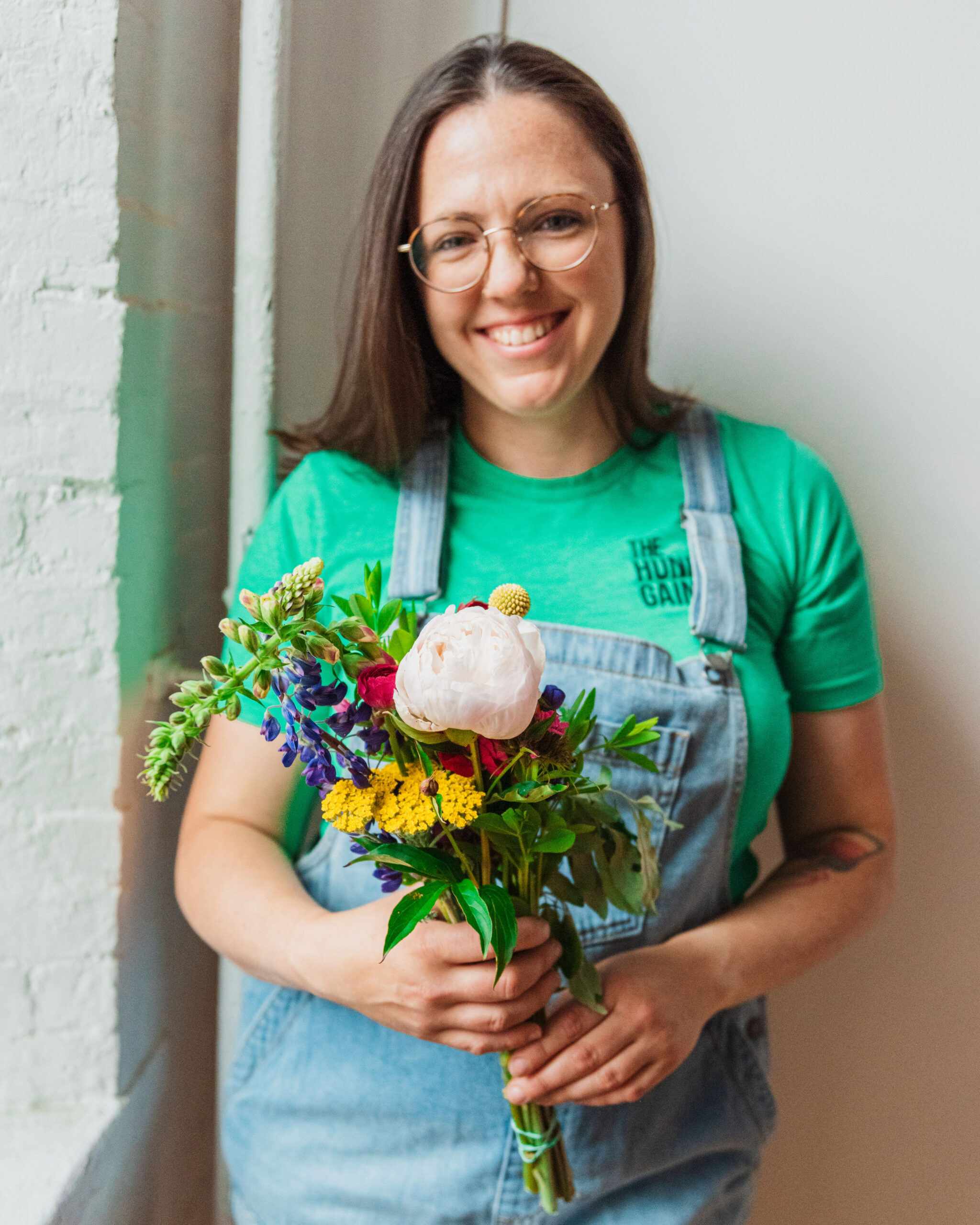 Alicia Valcaniant in overalls and green t-shirt holding a bouquet of flowers and smiling