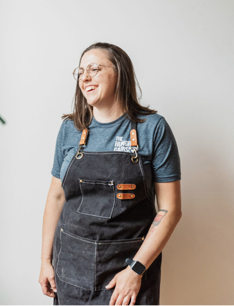 Hungry Gains owner Alicia Valcaniant wearing a black apron and a blue t-shirt, laughing against a white background.